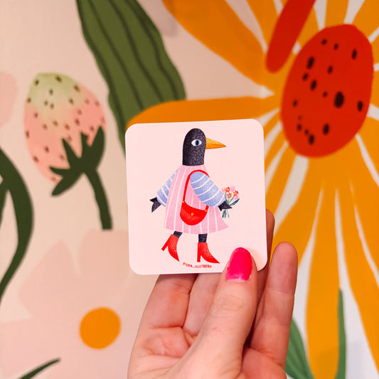 A hand with pink nail polish holds a square art print featuring a stylized penguin in a pink dress and red boots, set against a colorful floral background.