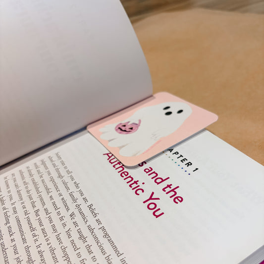 A pink bookmark with a cute cartoon ghost and pumpkin design rests on an open book page with red text reading 'Chapter 1: and the Authentic You'.