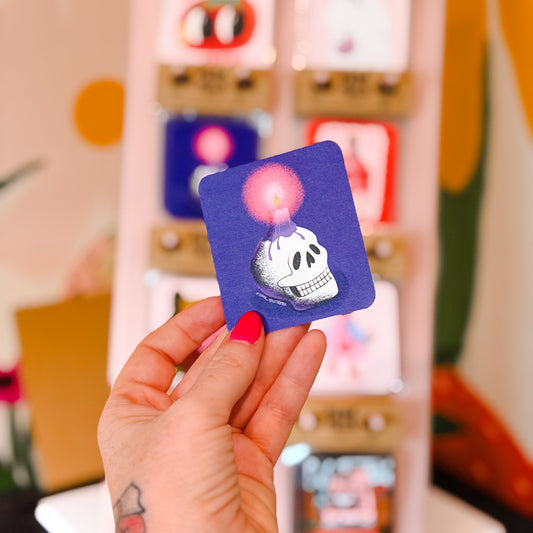 A hand with pink nail polish holds a small square purple sticker featuring a white skull and a glowing pink bubble. In the blurred background, a display stand shows other colorful stickers.