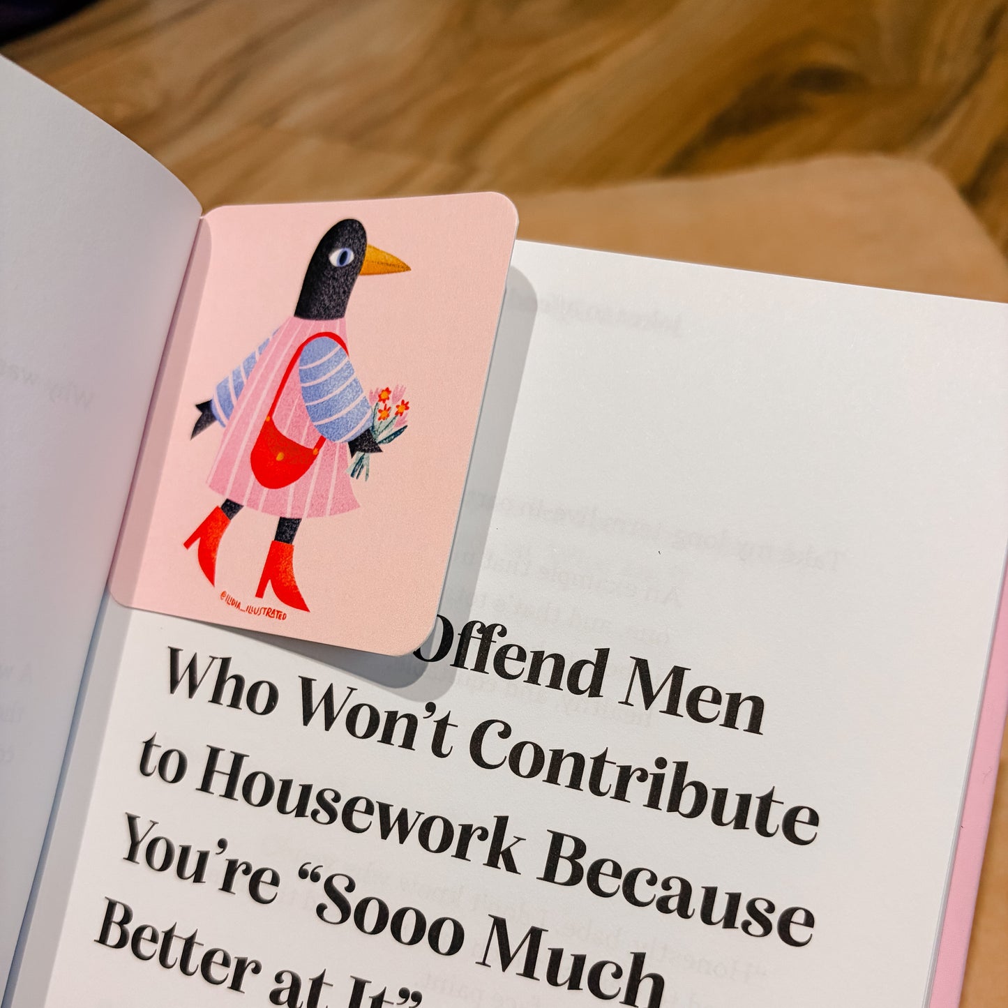 A pink rectangular book bookmark featuring a cartoon penguin in a pink dress and red boots, placed on an open book with a humorous housework quote printed on the page.