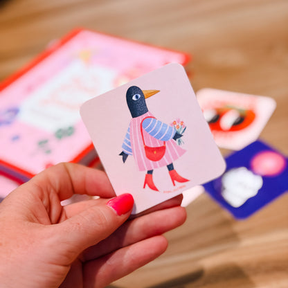 A hand with bright pink nail polish holds a square card featuring a stylized penguin in a pink and blue outfit. Other colorful cards are blurred in the background on a wooden surface.