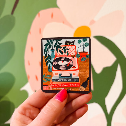 A hand with bright pink nail polish holds a square enamel pin featuring a retro illustration of a record player, cat, and plants. The background is a colorful, out-of-focus floral mural.