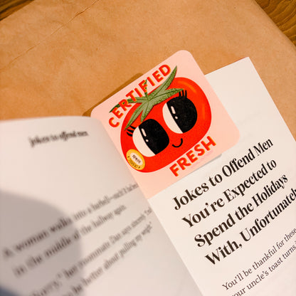 A square sticker featuring a cartoon tomato with the words 'CERTIFIED FRESH' is placed on top of an open printed booklet on a brown kraft-paper surface.