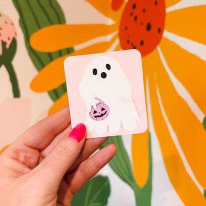 A hand with pink nail polish holds a square sticker featuring a white cartoon ghost holding a small jack-o'-lantern candy bucket, set against a colorful floral background.