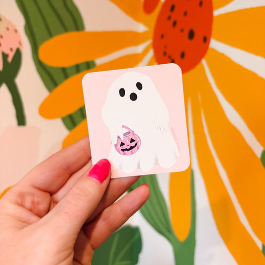 A hand with pink nail polish holds a square sticker featuring a white cartoon ghost holding a small jack-o'-lantern candy bucket, set against a colorful floral background.