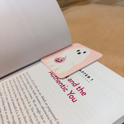 A pink bookmark with a cute cartoon ghost and pumpkin design rests on an open book page with red text reading 'Chapter 1: and the Authentic You'.