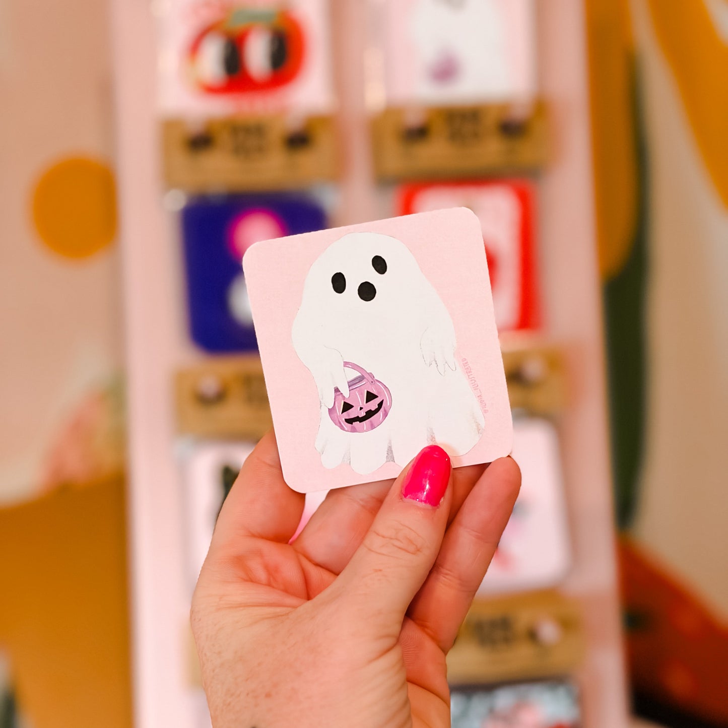 A hand with bright pink nail polish holds a small square sticker featuring a white cartoon ghost holding a jack-o'-lantern. In the blurred background, a display box contains other colorful stickers.