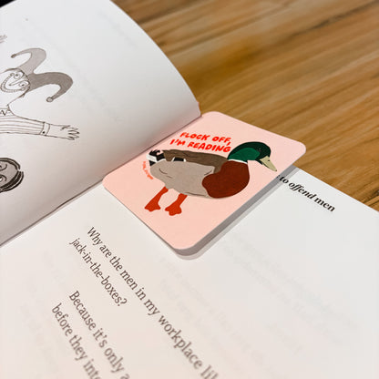 A rectangular pink book bookmark with a cartoon duck illustration and the text 'FLOCK OFF I'M READING' rests on an open book on a wooden table.