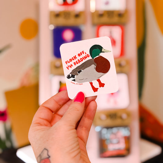 A hand with pink nail polish holds a square sticker featuring a cartoon mallard duck and the text 'CHECK OFF, I'M READING'. In the blurred background, a display box containing various colorful stickers is visible.