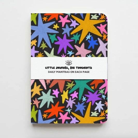A hardcover notebook with a colorful star pattern on a black background, featuring a white paper band around the middle with the text 'LITTLE JOURNEYS, BIG THOUGHTS' and 'DAILY MANTRAS ON EACH PAGE'.