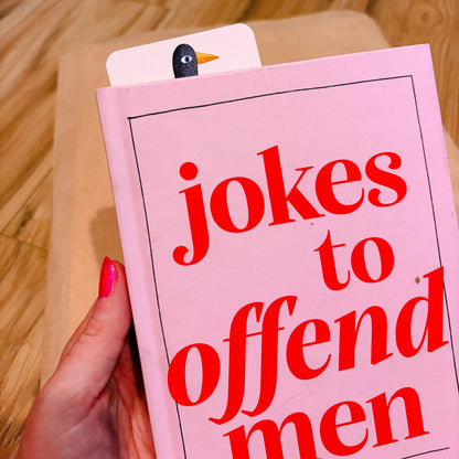 A hand with pink nail polish holds a pink book titled 'jokes to offend men' in a cozy indoor setting with a wooden floor background. A small penguin bookmark is visible at the top of the book.