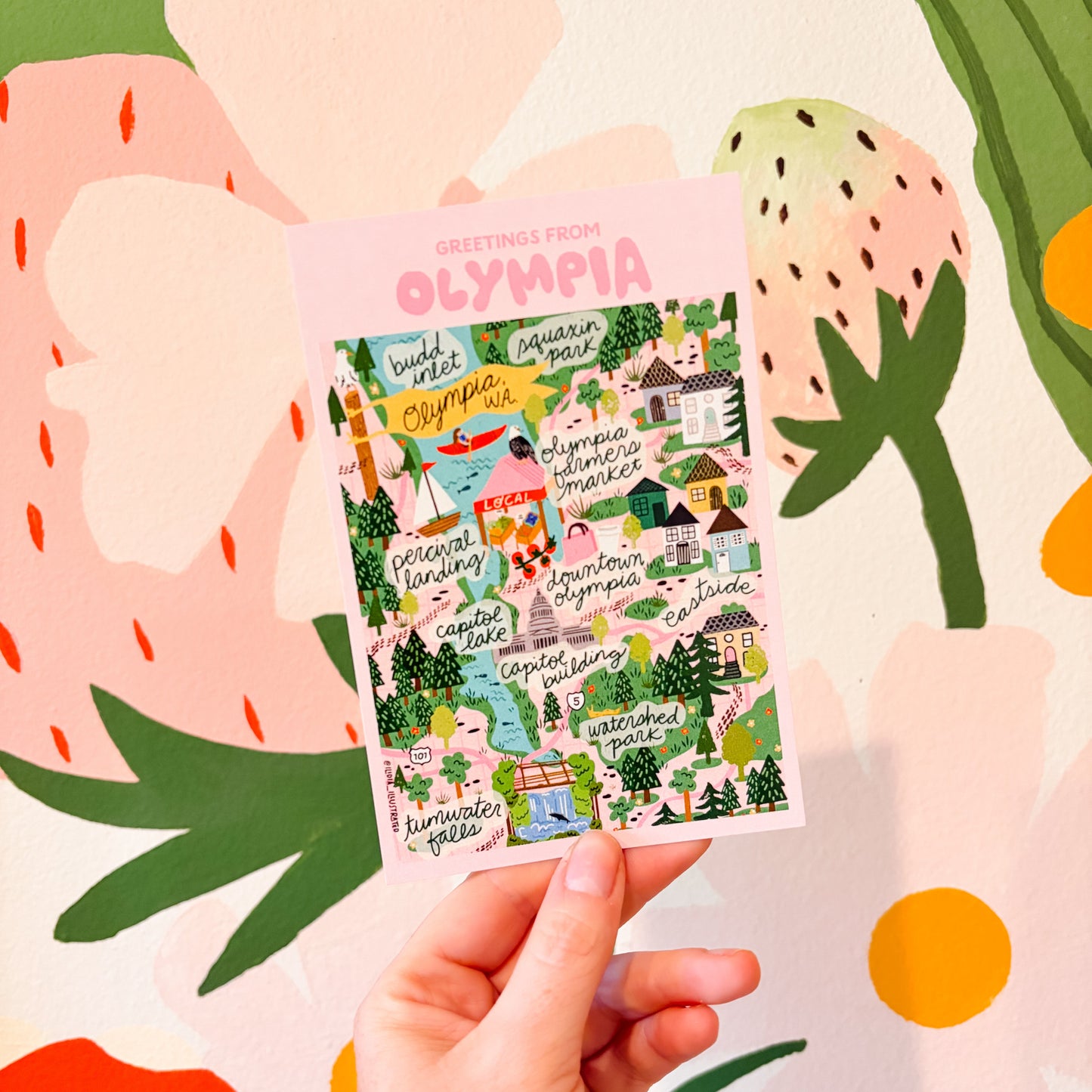 Hand holding a colorful Olympia map postcard 4x6 with 'Olympia' branding against a vibrant, abstract background.