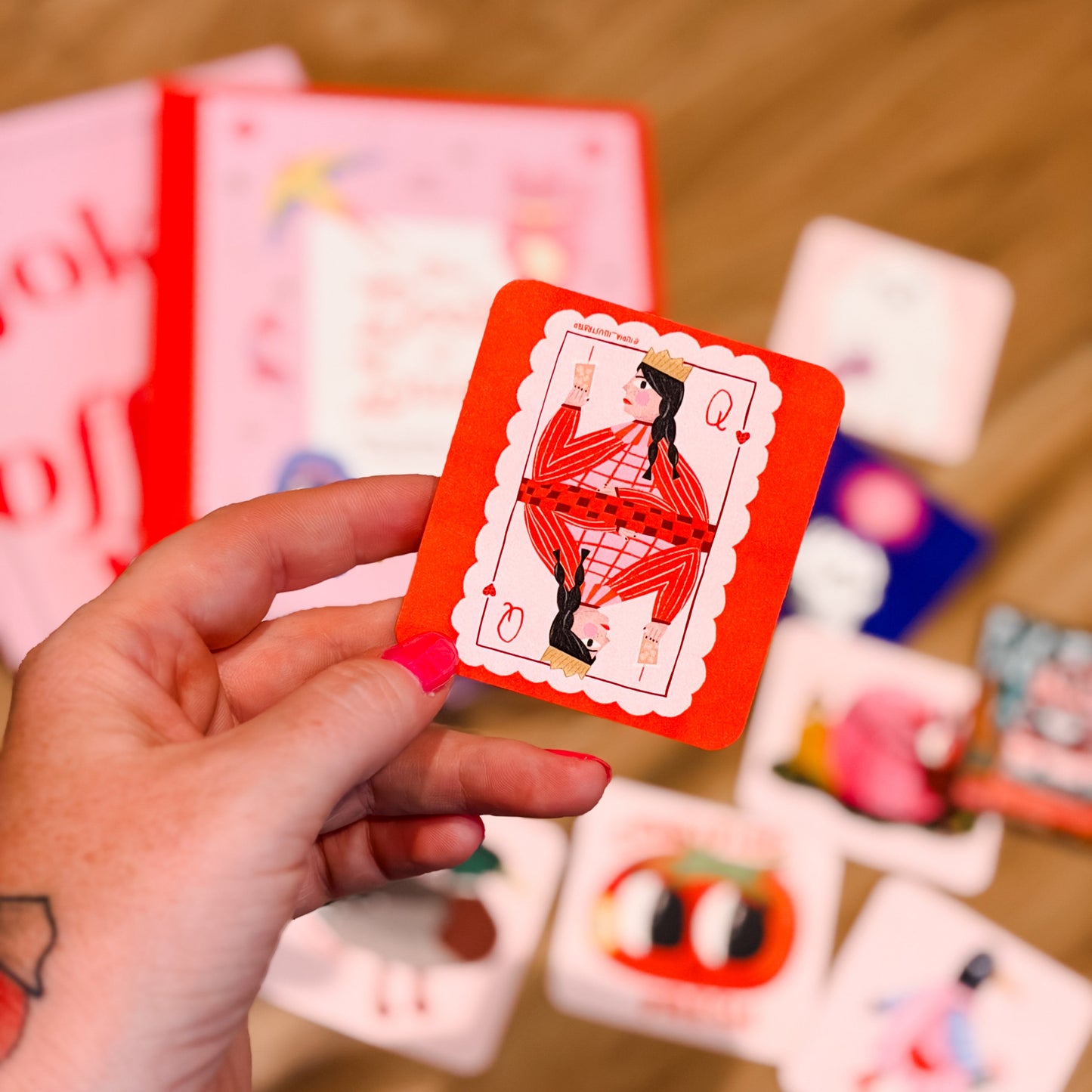 A hand holds a red playing card with a stylized figure illustration, with other colorful cards scattered on a wooden floor in the background.