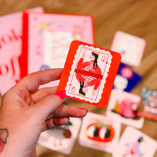 A hand holds a red playing card with a stylized figure illustration, with other colorful cards scattered on a wooden floor in the background.