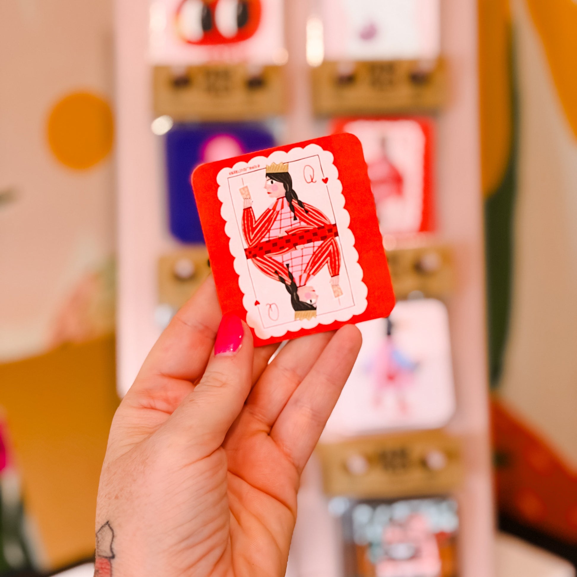 A hand with pink nail polish holds a small red and white illustrated card or tag in front of a blurred display of similar tags.