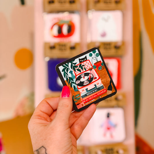 A hand with pink nail polish holds a small square illustrated card or sticker in front of a blurred display of similar colorful cards.