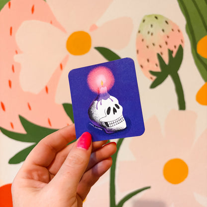 A hand with pink nail polish holds a square blue sticker featuring a white skull with a glowing pink candle on top, set against a colorful floral background.