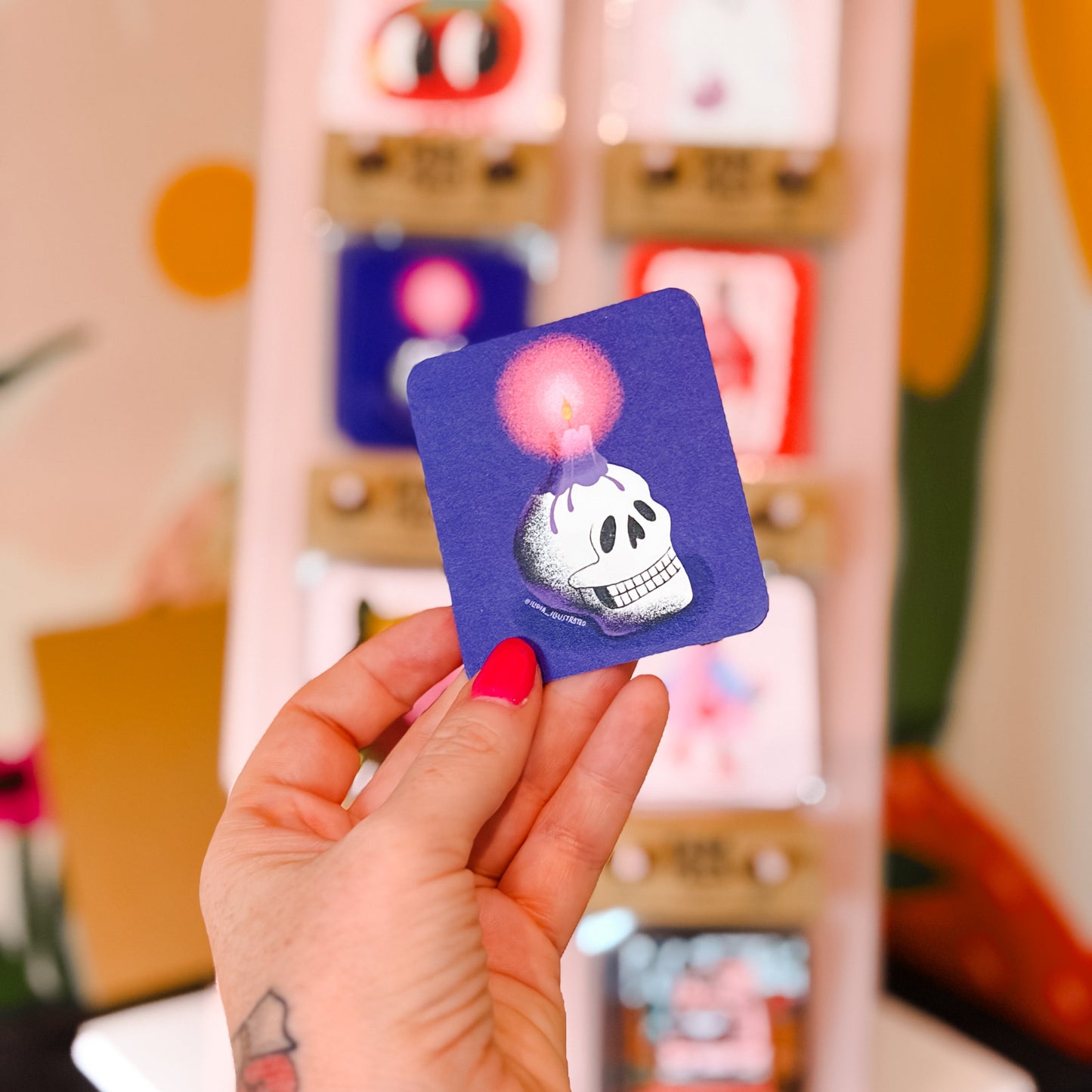 A hand with pink nail polish holds a small square purple sticker featuring a white skull and a glowing pink bubble. In the blurred background, a display stand shows other colorful stickers.