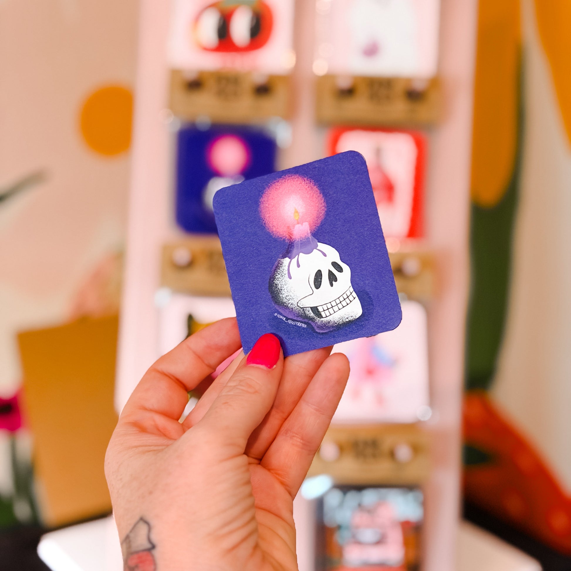 A hand with pink nail polish holds a small square purple sticker featuring a white skull and a glowing pink bubble. In the blurred background, a display stand shows other colorful stickers.