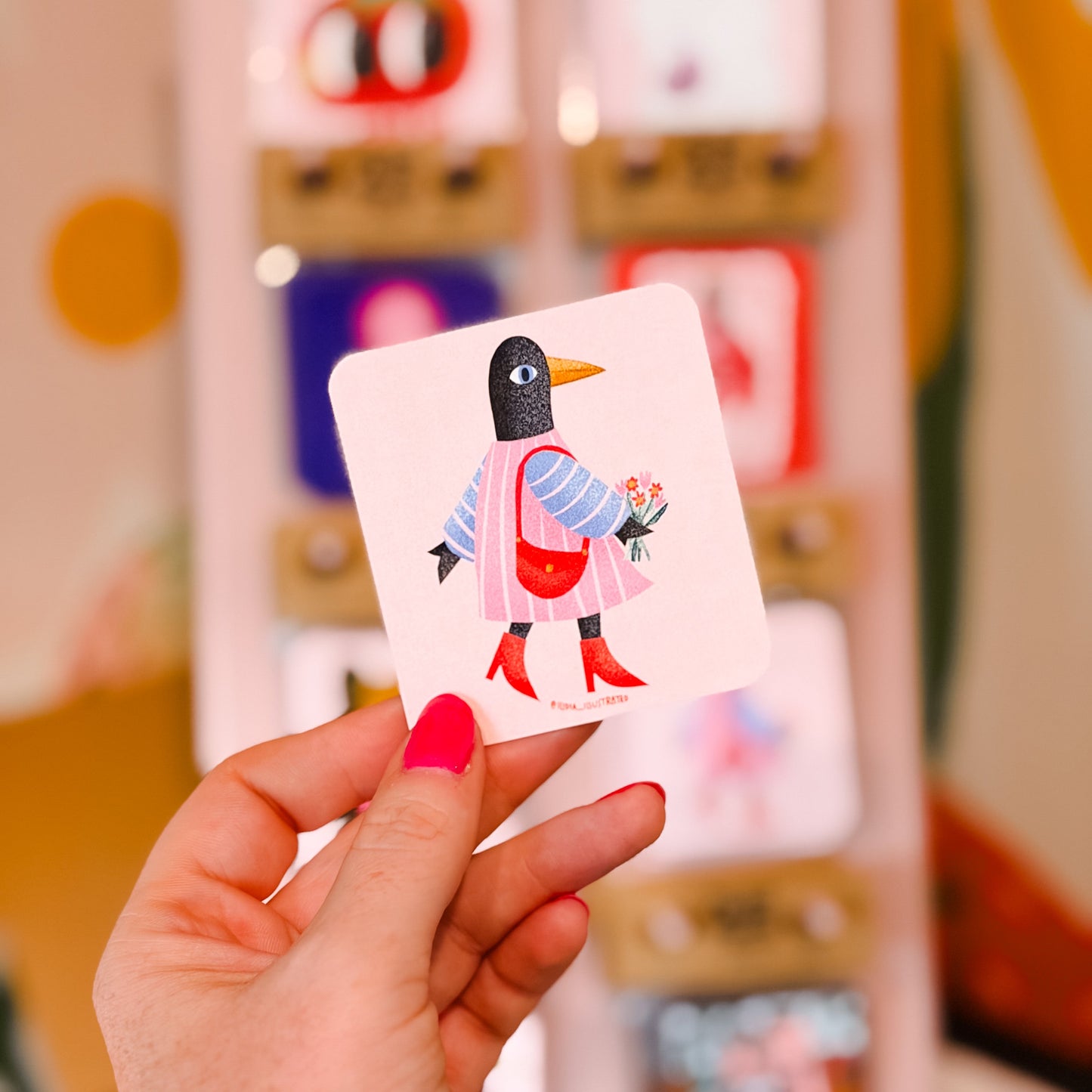 A hand with bright pink nail polish holds a square card featuring a stylized penguin in a pink dress and red boots, with a blurred display of similar cards in the background.