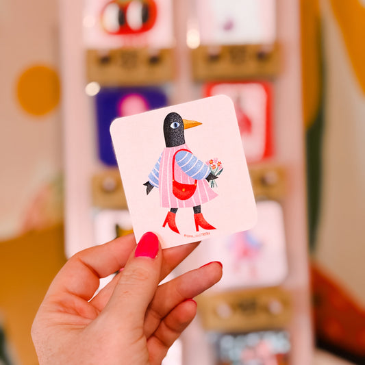 A hand with bright pink nail polish holds a square card featuring a stylized penguin in a pink dress and red boots, with a blurred display of similar cards in the background.