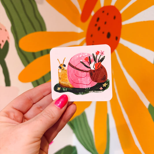 A hand with pink nail polish holds a small square card featuring a whimsical illustration of a pink snail and a yellow snail against a colorful floral background.