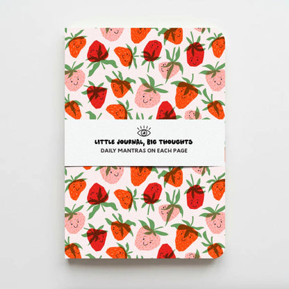 A notebook with a white cover featuring a repeating pattern of cute, smiling strawberries in red, pink, and orange with green leaves. A white paper band wrapped around the center reads 'LITTLE JOURNAL, BIG THOUGHTS' and 'DAILY MANTRAS ON EACH PAGE'.