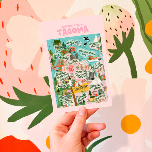 Hand holding a colorful postcard with 'Greetings from Tacoma' text against a floral background.