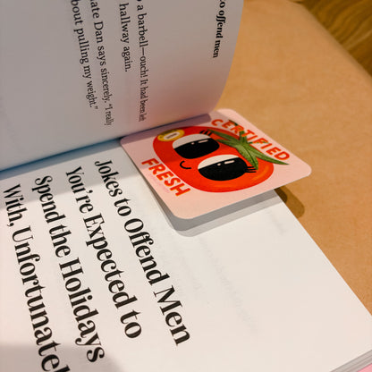 A square pink bookmark or card with a cartoon tomato face and the words 'CENSORED FRESH' resting on open book pages.