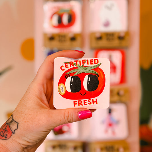 A hand with pink nail polish and tattoos holds a square white card featuring a red cartoon tomato face with the words 'CERTIFIED FRESH'. The background shows a blurred display of other colorful cards.