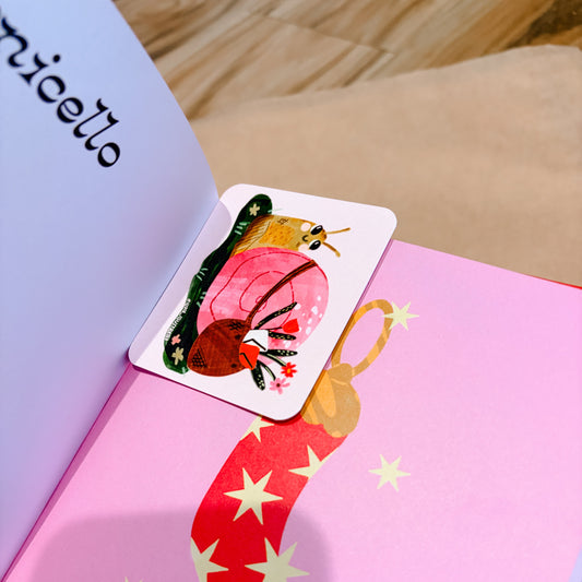 A small illustrated card or bookmark with a whimsical character sits on an open pink book. The book page shows a large red chili pepper with white stars, and part of another page is visible with the word 'micello' written on it.