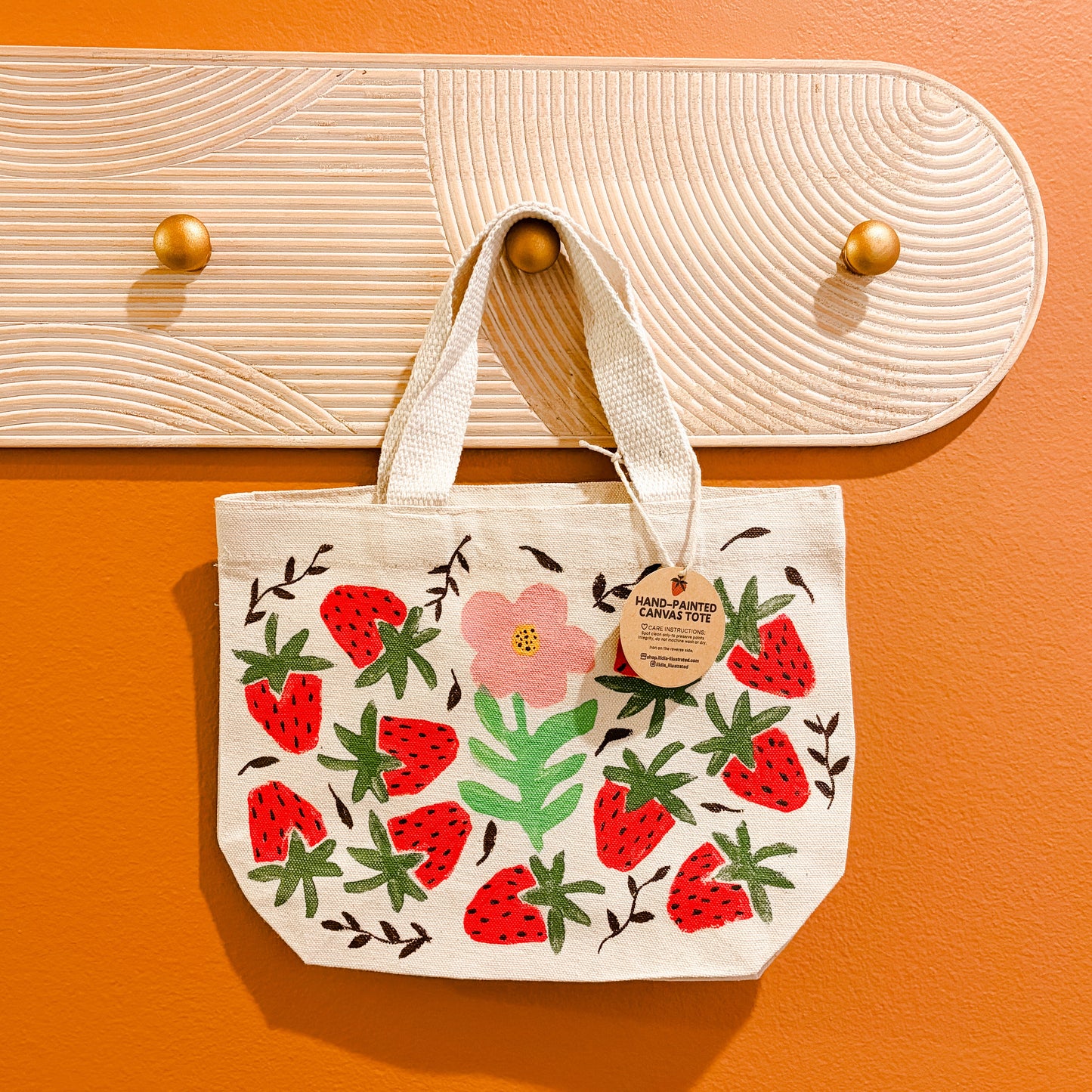 Tote bag with strawberry hand stamped design hanging on a wooden hook against an orange wall.
